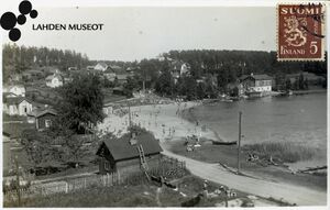 valokuvassa mustavalkoinen postikortti jossa valokuva uimarannasta ja rannalla ihmisiä. Rannan ympärillä asuinrakennuksia