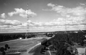 valokuvassa kaksi radiomastoa, radiomäen nurmikenttä ja taustalla tornien välissä tiilirakennus. Oikealla sivulla metsää.