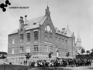 Lahden yhteiskoulu, etualalla koulun oppilaat, rehtori N. J. Avellan keskellä osittain pienen puun takana, kuvattu 1900-1903, kuvaaja Axel Strandberg. Lähde: Lahden kaupunginmuseon kuva-arkisto.